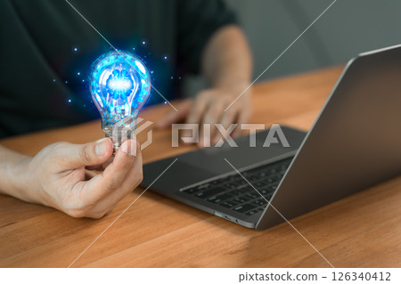 A person holding a glowing light bulb with a digital brain icon, symbolizing creativity, innovation, and ideas, next to a laptop on a wooden desk. 126340412