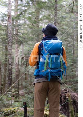 Back view of a faceless man carrying a backpack climbing a mountain and gazing at the fresh greenery 126340872
