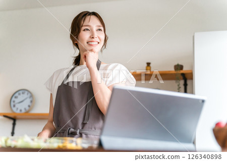 一位身穿圍裙的女士一邊做飯,一邊使用平板電腦上的應用程式查看食譜 一位身穿圍裙的女士一邊做飯,一邊使用平板電腦上的應用程式查看食譜 126340898