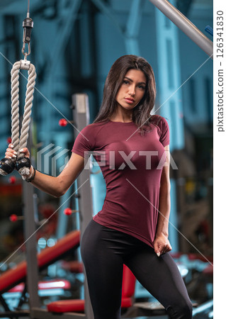 young woman in a maroon shirt confidently poses and holds fitness cables in a modern gym filled with equipment. She showcases her strength in an active environment. young woman in a maroon shirt confidently poses and holds fitness cables in a modern gym filled with equipment. She showcases her strength in an active environment. 126341830