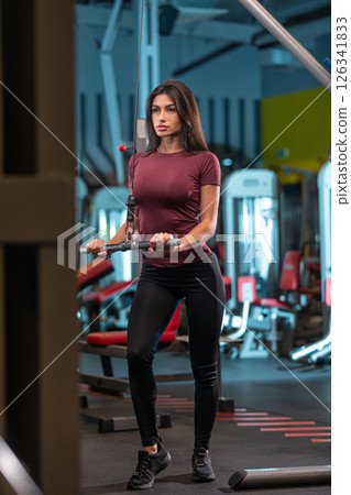 focused woman performs a strength training exercise using a cable machine in a well equipped gym. She wears a fitted top and leggings for optimal movement during her workout session. 126341833