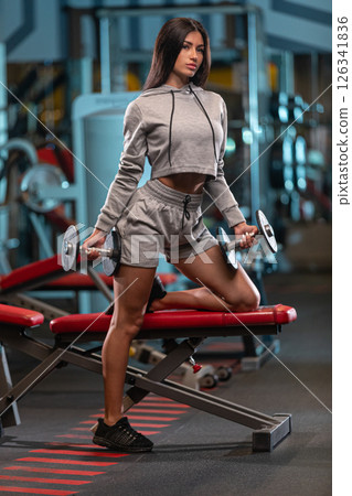 woman in athletic attire engaged in workout routine holding dumbbells while balancing on one knee on weight bench in well equipped gym early in day. 126341836