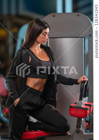 fitness enthusiast kneels beside a workout machine in a gym focusing intently while dressed in black athletic clothing. Evening light creates a motivating atmosphere. 126341843