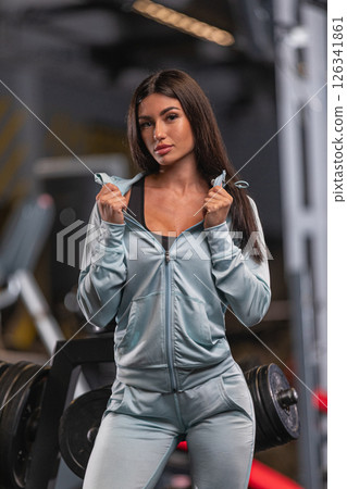 Young woman stands strong in a gym exuding confidence while wearing stylish athletic clothing. She poses with determination amid workout equipment designed for fitness training. 126341861