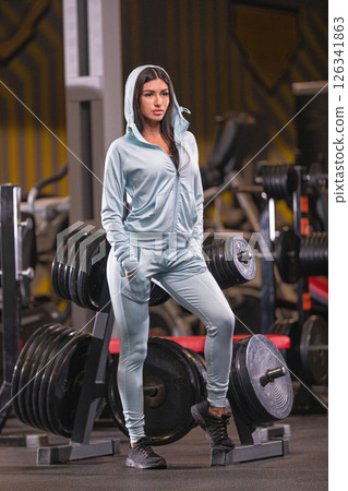 fit individual stands confidently in a gym dressed in activewear. The atmosphere is energized with weightlifting equipment around showcasing dedication to fitness and strength. 126341863