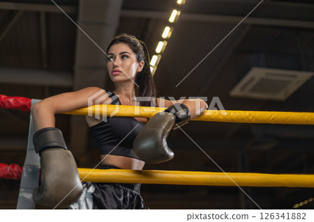 dedicated female boxer leans on ropes of boxing ring showcasing her strength and concentration while preparing for an intense training session in an indoor gym. 126341882
