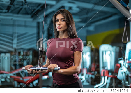 fitness enthusiast is performing strength training exercises on a cable machine in a well equipped gym. Focused on her form she displays determination. fitness enthusiast is performing strength training exercises on a cable machine in a well equipped gym. Focused on her form she displays determination. 126341914