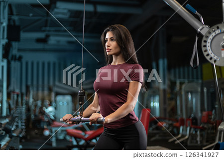 woman is focused on her workout as she uses a cable machine in a well equipped gym. The environment is spacious with various fitness equipment visible in the background. 126341927