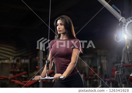 woman channels her energy as she trains with cable machine in modern gym showcasing her dedication to fitness and strength development during an evening workout. 126341928