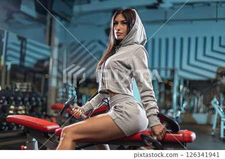 focused young woman wearing a gray hoodie and matching shorts lifts weights while sitting on a bench in a modern gym. The environment is equipped with various fitness tools. 126341941