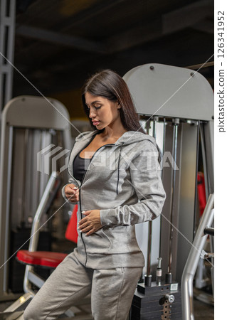 Fitness enthusiast in a gray activewear outfit stands beside gym equipment focusing on her workout routine in a modern gym environment. She maintains a determined expression. Fitness enthusiast in a gray activewear outfit stands beside gym equipment focusing on her workout routine in a modern gym environment. She maintains a determined expression. 126341952