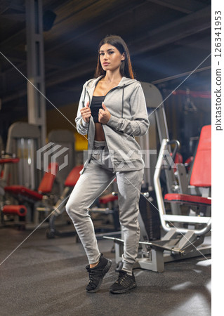 woman stands in fitness attire in a gym showcasing her dedication to health and fitness. She is surrounded by gym equipment and displays a strong posture ready to exercise. woman stands in fitness attire in a gym showcasing her dedication to health and fitness. She is surrounded by gym equipment and displays a strong posture ready to exercise. 126341953