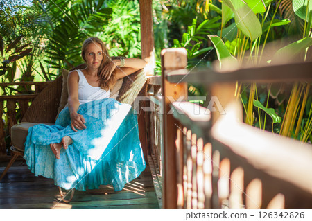 Young woman sit on open-air veranda of wooden bungalow with tropical garden view. Girl relaxing in luxury villa on family summer vacation. Countryside lifestyle. Young woman sit on open-air veranda of wooden bungalow with tropical garden view. Girl relaxing in luxury villa on family summer vacation. Countryside lifestyle. 126342826