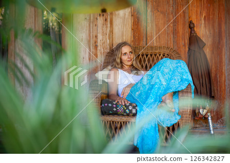 Young woman sit on open-air veranda of wooden bungalow with tropical garden view. Girl relaxing in luxury villa on family summer vacation. Countryside lifestyle. Young woman sit on open-air veranda of wooden bungalow with tropical garden view. Girl relaxing in luxury villa on family summer vacation. Countryside lifestyle. 126342827