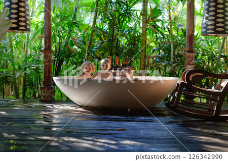 Happy babies have fun in bathtub, in outside bathroom on veranda with beautiful tropical garden view at luxury villa on summer family vacation. Happy babies have fun in bathtub, in outside bathroom on veranda with beautiful tropical garden view at luxury villa on summer family vacation. 126342990