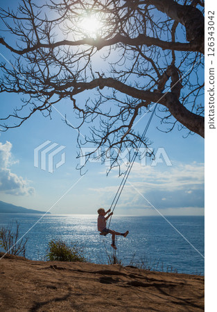 Happy girl have fun swinging high in mid air. Flying up upside down on rope swing on sea beach. Travel adventure on paradise tropical island. Family lifestyle, activity on summer vacation with kids. 126343042