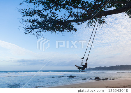 Happy girl have fun swinging high in mid air. Flying on rope swing on sea beach. Travel adventure on paradise tropical island. Family lifestyle, activity on summer vacation. 126343048