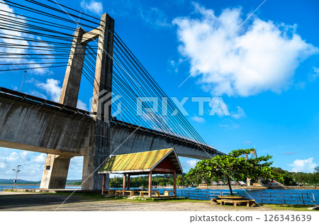 Koror - Babeldaob Japan - Palau Friendship Bridge under a bright blue sky. The modern cable-stayed structure spans a calm turquoise channel, with a shaded seating area, coastal trees and traditional Koror - Babeldaob Japan - Palau Friendship Bridge under a bright blue sky. The modern cable-stayed structure spans a calm turquoise channel, with a shaded seating area, coastal trees and traditional 126343639