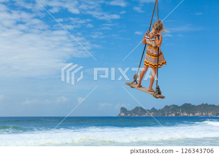 Happy girl have fun swinging high in mid air. Flying up upside down on rope swing on sea beach. Travel adventure on paradise tropical island. Family lifestyle, activity on summer vacation with kids. Happy girl have fun swinging high in mid air. Flying up upside down on rope swing on sea beach. Travel adventure on paradise tropical island. Family lifestyle, activity on summer vacation with kids. 126343706