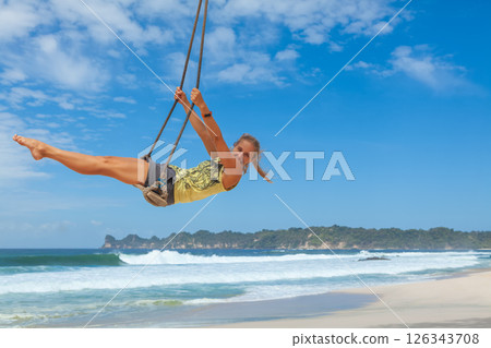 Happy girl have fun swinging high in mid air. Flying on rope swing on sea beach. Travel adventure on paradise tropical island. Family lifestyle, activity on summer vacation. Happy girl have fun swinging high in mid air. Flying on rope swing on sea beach. Travel adventure on paradise tropical island. Family lifestyle, activity on summer vacation. 126343708