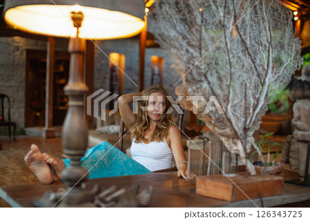 Young woman sit on sofa inside luxury wooden villa. Girl relaxing on family summer vacation. Tropical lifestyle. 126343725