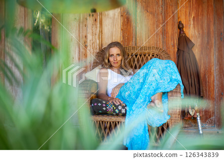 Young woman sit on open-air veranda of wooden bungalow with tropical garden view. Girl relaxing in luxury villa on family summer vacation. Countryside lifestyle. 126343839