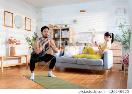 Young asian man doing squat exercise on yoga mat in living room at home while woman using phone. Young asian man doing squat exercise on yoga mat in living room at home while woman using phone. 126346690
