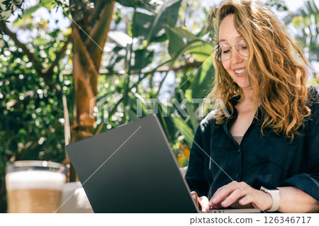 Smiling Woman Typing on Laptop While Working Outdoors in Tropical Garden with Coffee 126346717