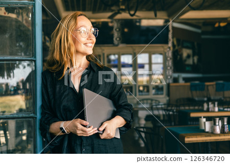 Smiling Woman with Laptop Standing by Cafe Window, Wearing Glasses and Smartwatch 126346720