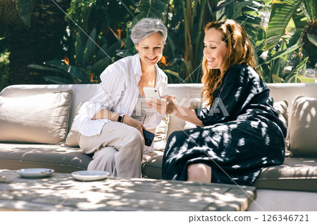 Two mature women smiling and looking at smartphone together while sitting outdoors on a terrace 126346721