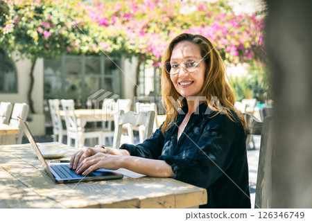 Smiling woman in glasses working on laptop at outdoor cafe surrounded by flowers on a sunny day 126346749
