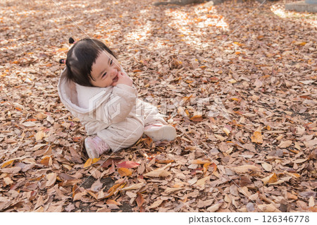 [One-year-old child sitting and playing in a winter park covered with fallen leaves] 126346778