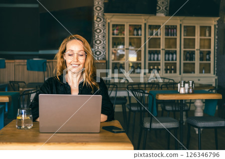Smiling woman using laptop in a trendy cafe, enjoying remote work with a drink. Cozy workspace  126346796