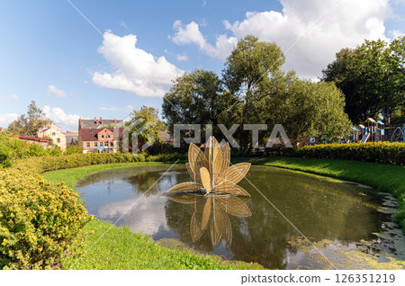 A peaceful pond features a lotus fountain amid lush greenery on a sunny day. A peaceful pond features a lotus fountain amid lush greenery on a sunny day. 126351219