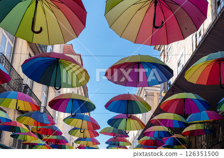 Brightly colored umbrellas are suspended above a lively street under a clear blue sky. Brightly colored umbrellas are suspended above a lively street under a clear blue sky. 126351500