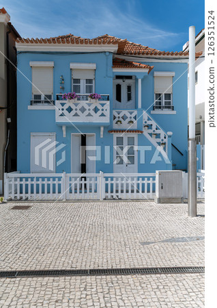 Bright blue beach house with balcony and white fence in a coastal town under clear sky Bright blue beach house with balcony and white fence in a coastal town under clear sky 126351524