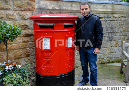 Man standing next to a bright red Royal Mail post box in Windsor, England, a traditional British symbol seen across the UK, during a sightseeing day. 126351575