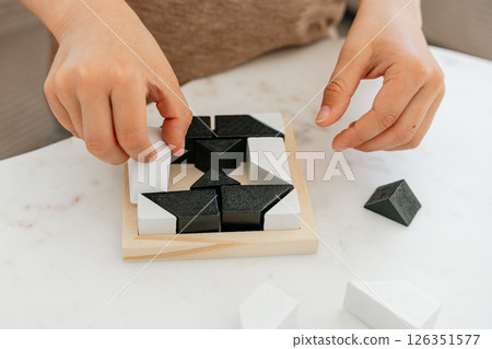 Hands placing black and white geometric puzzle pieces into a wooden frame. Close-up image showing the intricate task of arranging shapes to fit perfectly on a white surface. 126351577