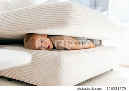 Child playfully hides between the cushions of a white sofa, displaying a curious and mischievous expression. The cozy setting suggests a fun and imaginative moment of play. 126351579