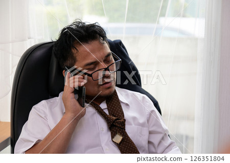 Disheveled businessman wearing glasses and loose tie sitting in office chair while talking on smartphone, appearing tired and stressed with window light in background 126351804