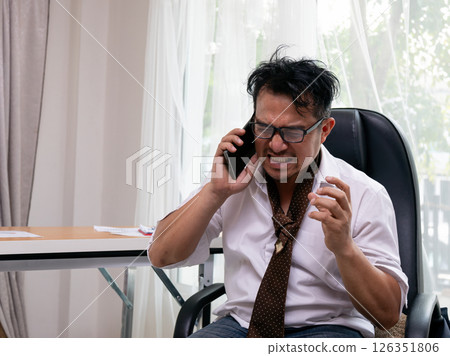 Angry businessman shouting during phone call with furious facial expression and clenched hand, sitting in office chair wearing disheveled white shirt and loose tie Angry businessman shouting during phone call with furious facial expression and clenched hand, sitting in office chair wearing disheveled white shirt and loose tie 126351806