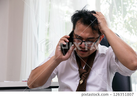 Stressed businessman holding head in frustration while talking on smartphone, wearing glasses and loose tie, sitting in office chair dealing with overwhelming work problems or bad news Stressed businessman holding head in frustration while talking on smartphone, wearing glasses and loose tie, sitting in office chair dealing with overwhelming work problems or bad news 126351807