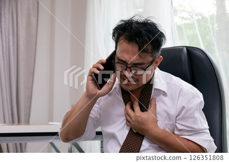 Frustrated businessman gripping chest in anxiety while talking on phone, stressed expression with wrinkled face, wearing glasses and loosened tie, sitting indoors in office Frustrated businessman gripping chest in anxiety while talking on phone, stressed expression with wrinkled face, wearing glasses and loosened tie, sitting indoors in office 126351808