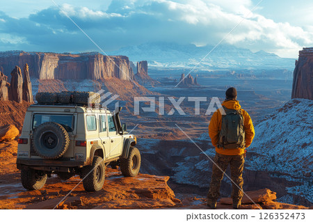 Off-road expedition truck parked on rocky plateau, adventurer standing beside it, gazing at vast canyons and towering mesas, capturing the raw beauty of untamed wilderness 126352473