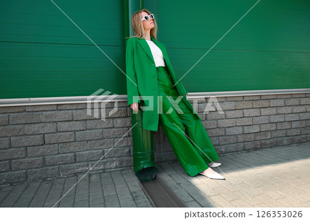 High fashion photo of a young blond woman in green coat and leather pants. Monochrome green outfit, white glasses and shoes. High fashion photo of a young blond woman in green coat and leather pants. Monochrome green outfit, white glasses and shoes. 126353026