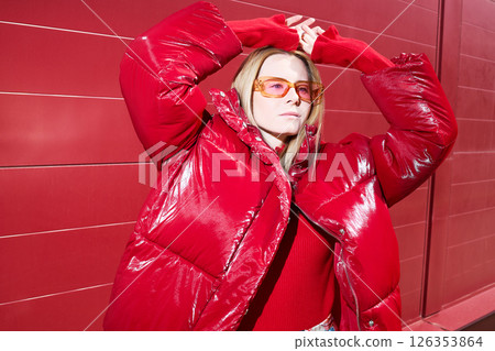 High fashion photo of a young blond woman in red glossy down jacket, poloneck, and pants. Monochrome trendy red outfit. 126353864