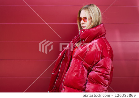 High fashion photo of a young blond woman in red glossy down jacket, poloneck, and pants. Monochrome trendy red outfit. High fashion photo of a young blond woman in red glossy down jacket, poloneck, and pants. Monochrome trendy red outfit. 126353869