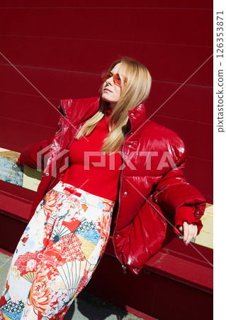 High fashion photo of a young blond woman in red glossy down jacket, poloneck, and pants. Monochrome trendy red outfit. 126353871