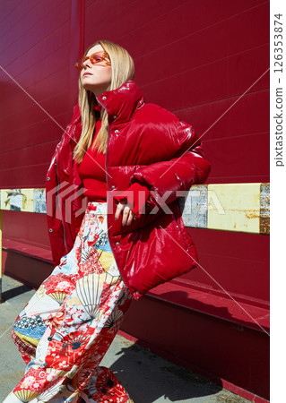 High fashion photo of a young blond woman in red glossy down jacket, poloneck, and pants. Monochrome trendy red outfit. 126353874