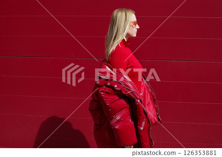 High fashion photo of a young blond woman in red glossy down jacket, poloneck, and pants. Monochrome trendy red outfit. 126353881